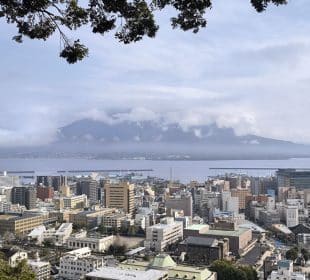 Eine Stadtlandschaft mit vielen Gebäuden am Wasser, eingerahmt von grünen Zweigen, mit dem Vulkan Sakurajima im Hintergrund, der teilweise von Wolken bedeckt ist - aufgenommen von der Aussichtsplattform des Shiroyama-Parks in Kagoshima.