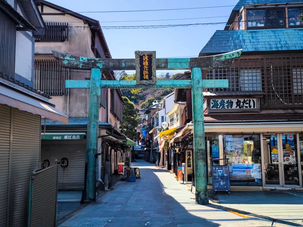 Ein verwittertes grünes Torii-Tor, bekannt als Hetsunomiya, steht am Eingang einer schmalen Straße mit traditionellen japanischen Geschäften unter einem klaren blauen Himmel. Über einigen Geschäften hängen Schilder mit japanischer Schrift.