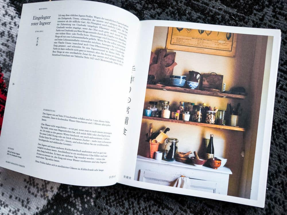Ein aufgeschlagenes Kochbuch auf einem gemusterten Stoff zeigt links eine Textseite und rechts ein Foto eines rustikalen Küchenregals mit Gläsern, Schüsseln und Utensilien. Die Holzregale zeigen verschiedene Küchenutensilien und weisen auf Geschichte und Traditionen wie Gohan hin.