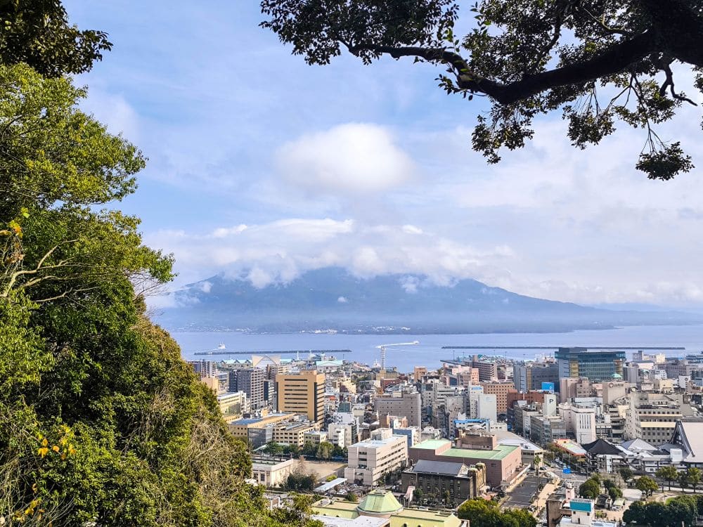 Von der Aussichtsplattform des Shiroyama-Parks blickt man auf eine Küstenstadt mit hohen Gebäuden an einem mit Bäumen bewachsenen Hang. Im Hintergrund erhebt sich der Vulkan Sakurajima bei teilweise bewölktem Himmel über das Wasser.