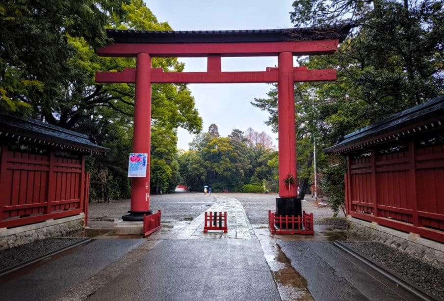 Ein großes rotes Torii-Tor steht am Eingang des Hikawa-Schreins, flankiert von roten Holzzäunen und umgeben von üppig grünen Bäumen an einem nassen, bewölkten Tag in Saitama.
