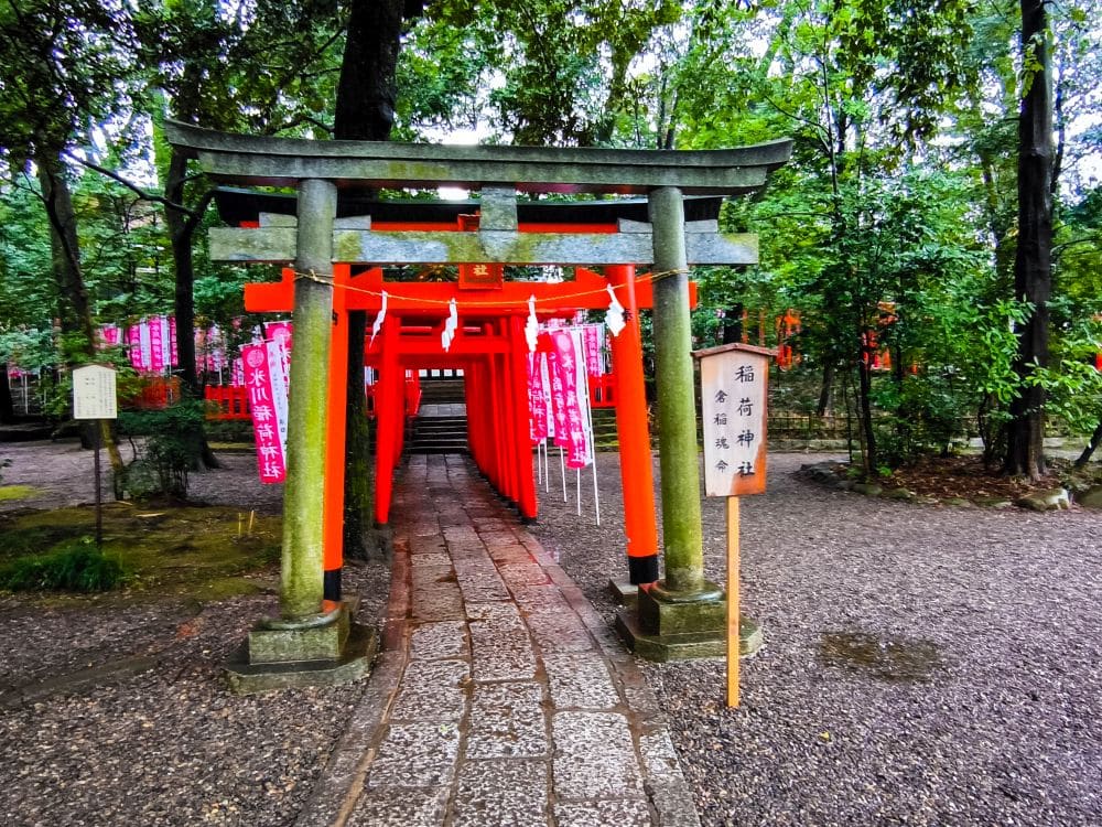 Ein mit mehreren leuchtend roten Torii-Toren gesäumter Weg führt durch ein üppiges, grünes Waldgebiet in Japan. Ein steinerner Wegweiser mit japanischer Schrift steht am Eingang neben den Toren und zeigt den Eingang zum Hikawa-Schrein an. Der Boden ist mit Schotter bedeckt.