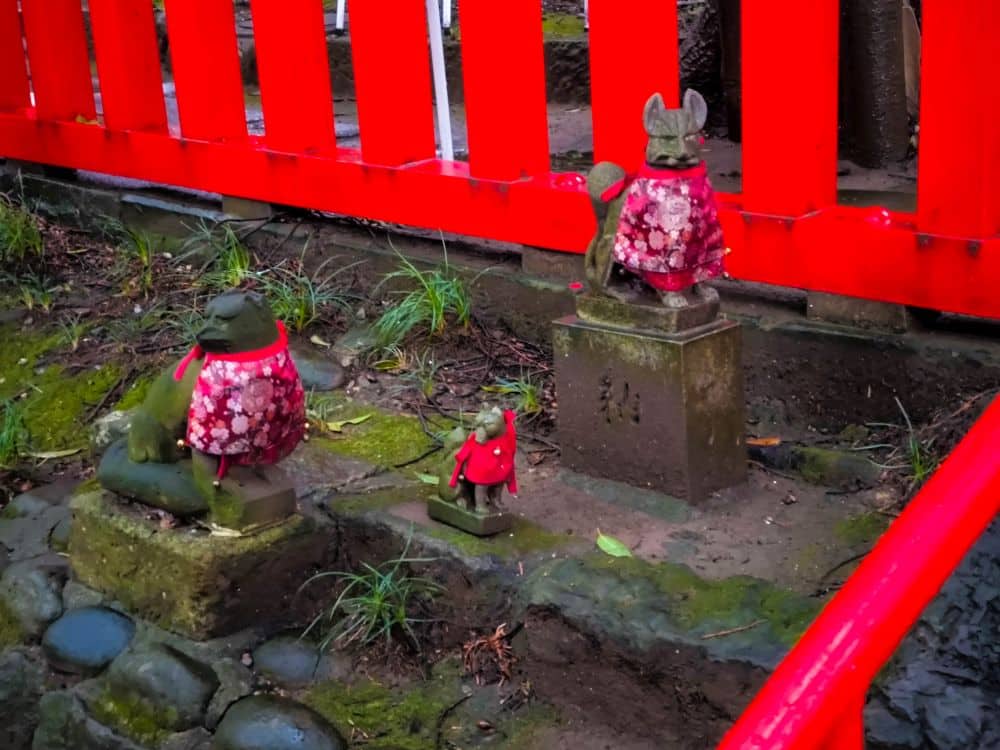 Drei Tierfiguren, gekleidet in rote Tücher mit weißen Mustern, sitzen auf Steinen und kleinen Plattformen neben einem leuchtend roten Hikawa-Zaun. Der Boden ist schlammig und mit Gras- und Moosflecken übersät.