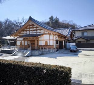 Ein traditionelles japanisches Gebäude mit Holzrahmen und Ziegeldach, wahrscheinlich Teil des Hosenji-Tempels in Hino, steht neben einem modernen Haus. Ein schwarzes Auto ist in der Nähe geparkt und Bäume umrahmen diesen Ort der Ruhe unter einem klaren blauen Himmel.