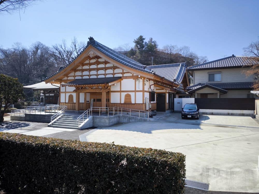 Ein traditionelles japanisches Gebäude mit Holzrahmen und Ziegeldach, wahrscheinlich Teil des Hosenji-Tempels in Hino, steht neben einem modernen Haus. Ein schwarzes Auto ist in der Nähe geparkt und Bäume umrahmen diesen Ort der Ruhe unter einem klaren blauen Himmel.