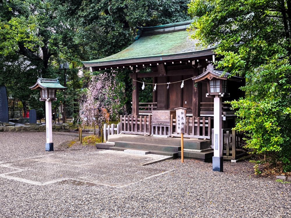 Ein kleiner traditioneller japanischer Hikawa-Schrein mit grünem Dach und Holzkonstruktion, umgeben von Grün und Steinlaternen, steht in einem friedlichen Kieshof.