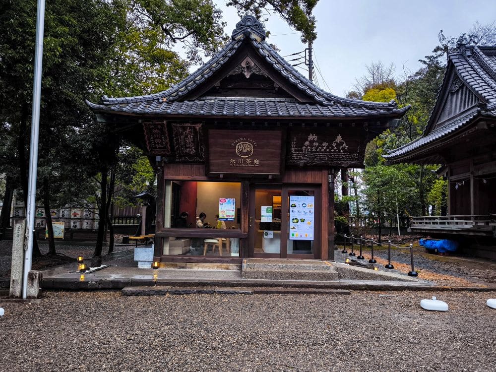 Ein kleiner Holzladen mit Ziegeldach in einem japanischen Garten, möglicherweise in der Nähe des Hikawa-Schreins, mit großen bunten Plakaten an der Fassade und warmen Lichtern entlang des Weges. Im Inneren sind durch ein Fenster Menschen zu sehen, und Bäume umgeben das Gebäude.