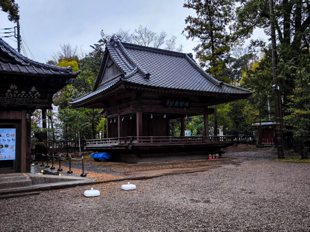 Ein traditionelles japanisches Holzgebäude, das möglicherweise mit Hikawa-Schreinen verbunden ist, steht an einem bewölkten Tag zwischen Bäumen, umgeben von Kies und verstreuten modernen Gegenständen. Auf der linken Seite sind ein Schild und kleine Schranken zu sehen.