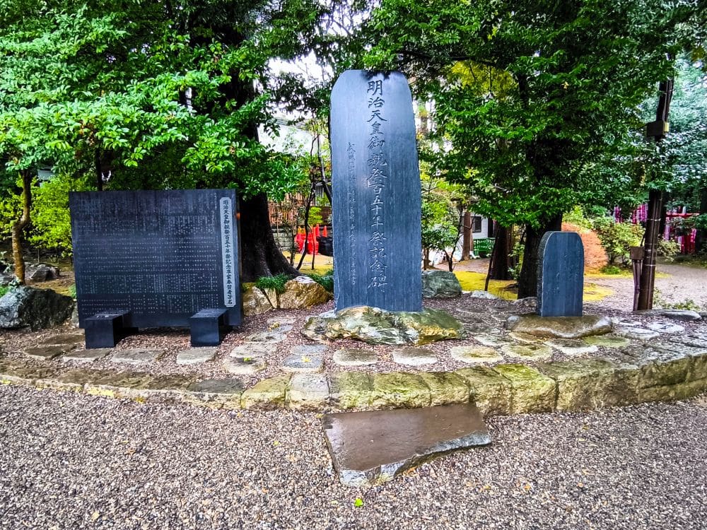 Steinmonumente mit japanischen Inschriften stehen umgeben von Bäumen und Kies in einer friedlichen Umgebung im Freien, wahrscheinlich an einer Gedenkstätte oder einem historischen Ort wie dem Hikawa-Schrein in Japan.