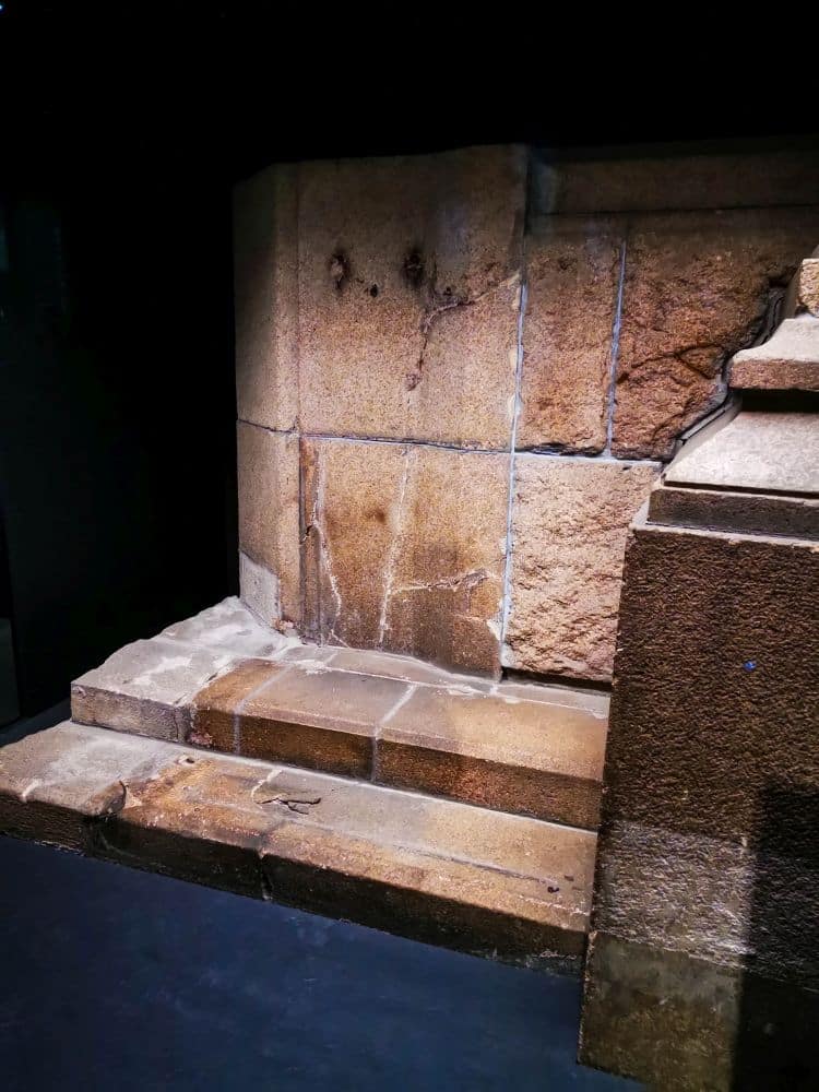 Große, verwitterte Steinblöcke bilden eine kleine, antike Treppe mit drei Stufen, die sich vor einem dunklen Hintergrund abzeichnet. Die abgenutzten Steine und die sichtbare Restaurierung deuten auf eine von Zeit und Frieden geprägte Geschichte hin.