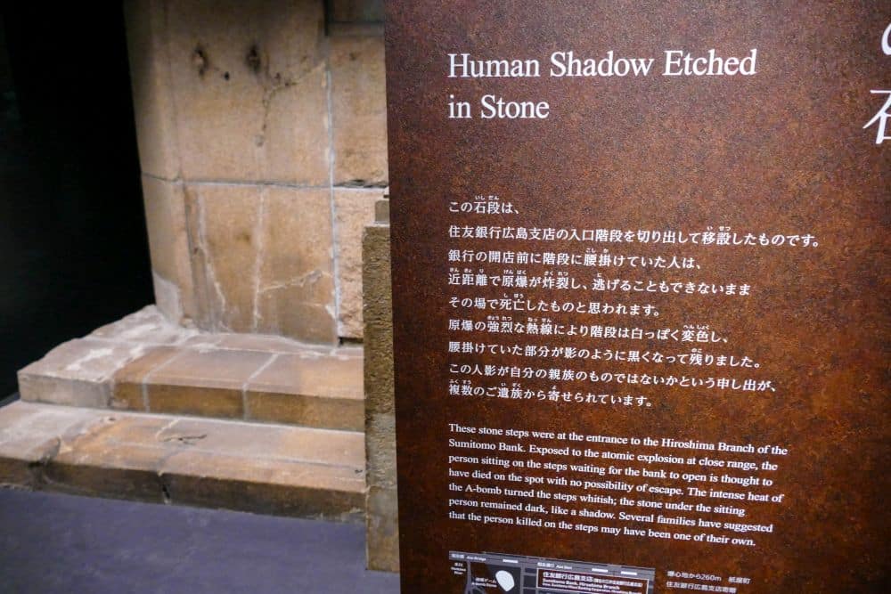 Auf einer Museumsanzeige ist ein Schild mit dem Titel "Human Shadow Etched in Stone" (Menschlicher Schatten in Stein gemeißelt) zu sehen, das eine steinerne Stufe beschreibt, die den Schatten der Hiroshima-Atombombe trägt und als eindringliche Erinnerung an den Frieden dient. Eine Treppe und eine Steinmauer stehen neben dem Schild.