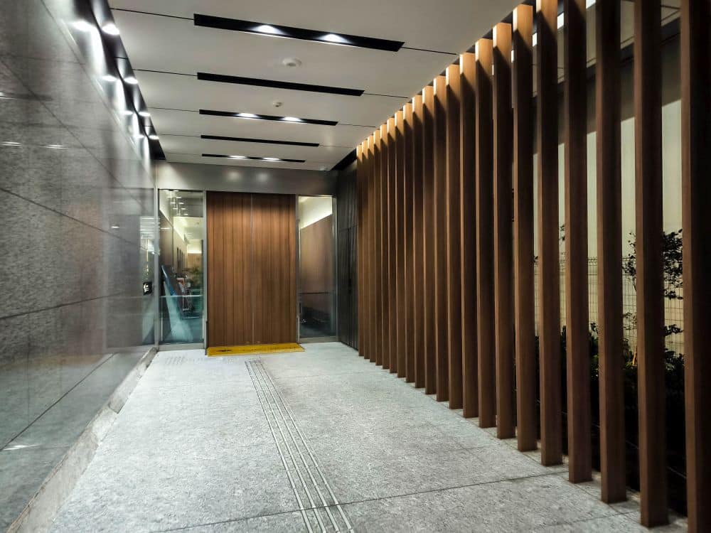 Ein moderner Flur mit einem von Tobu inspirierten Steinboden, vertikalen Holzlatten auf der einen Seite und glänzenden grauen Wandpaneelen auf der anderen. Deckenleuchten sorgen für eine schlichte, gut beleuchtete Umgebung. Am Ende befindet sich eine Holztür mit einem gelben taktilen Pflasterstreifen.