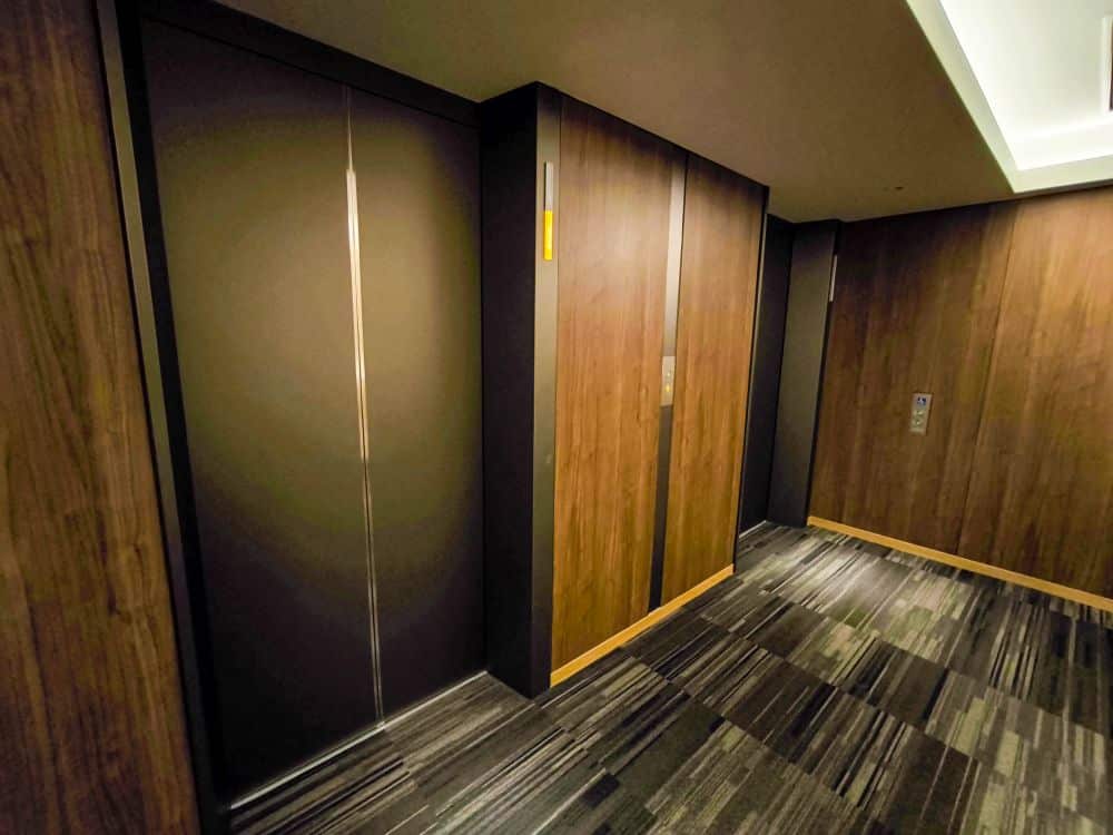 Zwei geschlossene Tobu-Aufzugstüren mit Holzverkleidung stehen in einem modernen Flur mit gemustertem Teppichboden und sanfter Deckenbeleuchtung.