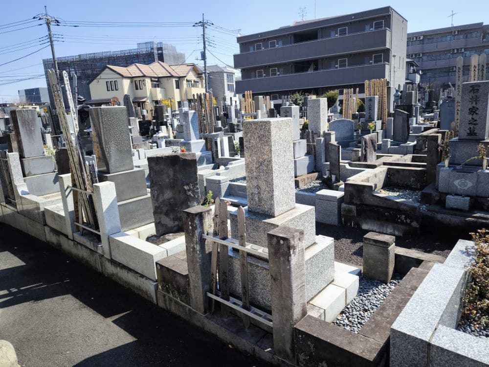 Reihen von grauen Steingrabsteinen auf dem Friedhof von Hosenji-Tempel, Hino, sind von städtischen Gebäuden und Stromleitungen unter einem klaren blauen Himmel umgeben. Einige Grabsteine sind mit hölzernen Gedenksteinen versehen, die die Geschichte dieses feierlichen Ortes widerspiegeln.