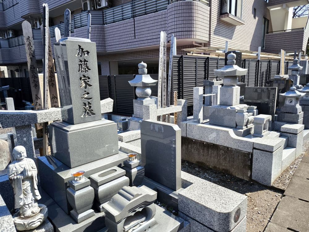 Auf dem Friedhof von Hosenji-Tempel, einem Ort der Ruhe in Hino, stehen Reihen von Grabsteinen und Denkmälern aus grauem Stein dicht beieinander; eine kleine Statue und Opfergaben sind zu sehen. Im Hintergrund erheben sich moderne Wohnhäuser.