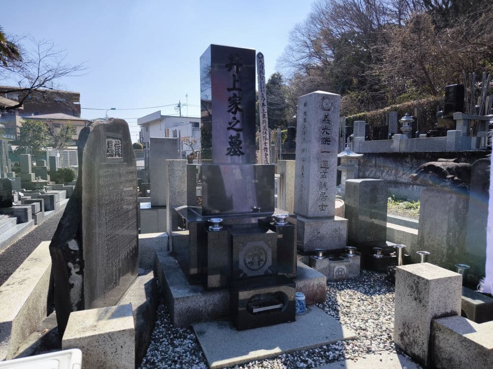 Auf dem japanischen Friedhof am Hosenji-Tempel in Hino befinden sich mehrere aufrecht stehende Grabsteine, darunter ein großer, polierter schwarzer Stein mit Kanji-Gravur. Dieser Ort der Ruhe ist von grauen Gräbern, Bäumen und entfernten Gebäuden umgeben.