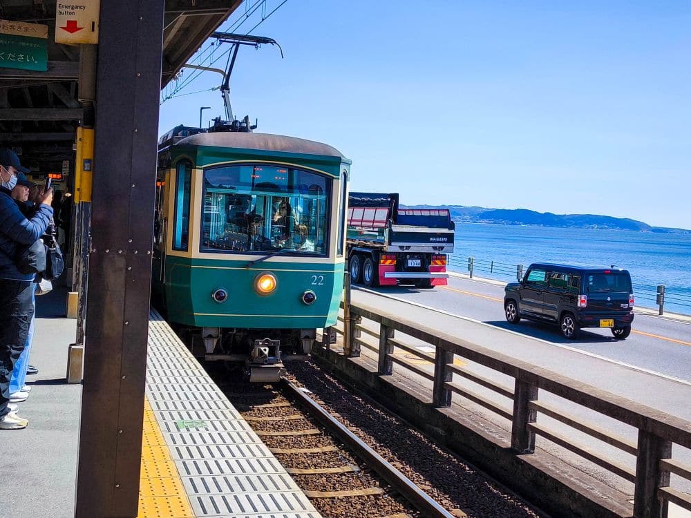 Ein grüner Zug fährt in den Bahnhof von Enoshima ein, wo Menschen auf dem Bahnsteig warten. Parallel zu den Gleisen verläuft eine Straße, auf der Autos und ein Lastwagen unter klarem Himmel am blauen Meer vorbeifahren.