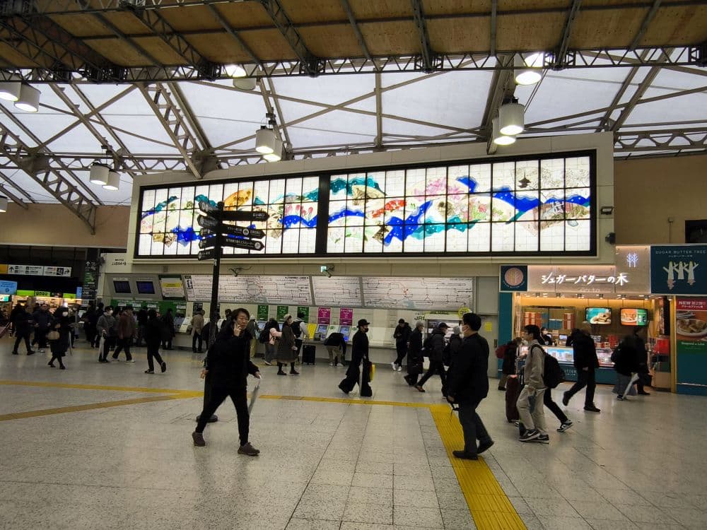 Die Menschen laufen durch den belebten Bahnhof von Hikawa mit seiner hohen Decke und dem großen Glasgemälde darüber. Es sind Fahrkartenautomaten, Schilder und Geschäfte zu sehen, und die meisten Menschen tragen dunkle Kleidung und Gesichtsmasken.