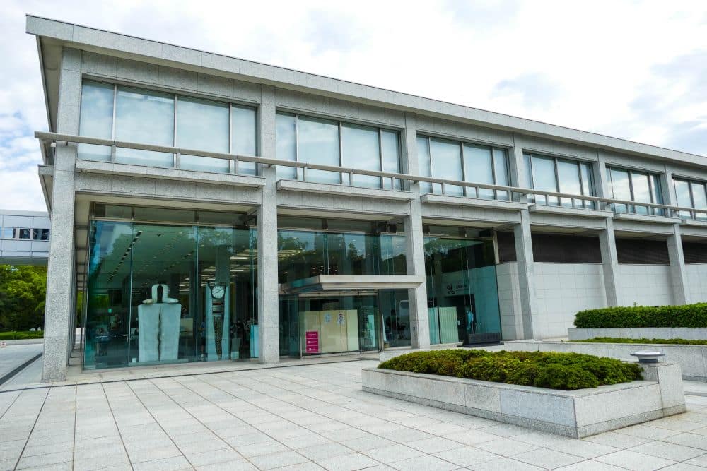 Modernes zweistöckiges Gebäude mit großen Glasfenstern und Steinfassade, in dem Schaufensterpuppen zu sehen sind. Der Eingang hat Glastüren, ein kleiner friedlicher Garten davor, und der Himmel ist teilweise bewölkt.