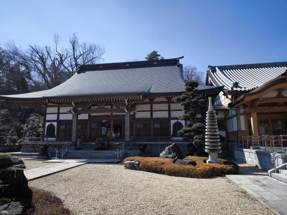 Der Hosenji-Tempel in Hino, einem Ort der Ruhe, erhebt sich mit geschwungenem Ziegeldach, Holzbalken und weißen Wänden in einem gepflegten Garten mit beschnittenen Büschen, Felsen und einer Steinpagode unter klarem blauem Himmel.