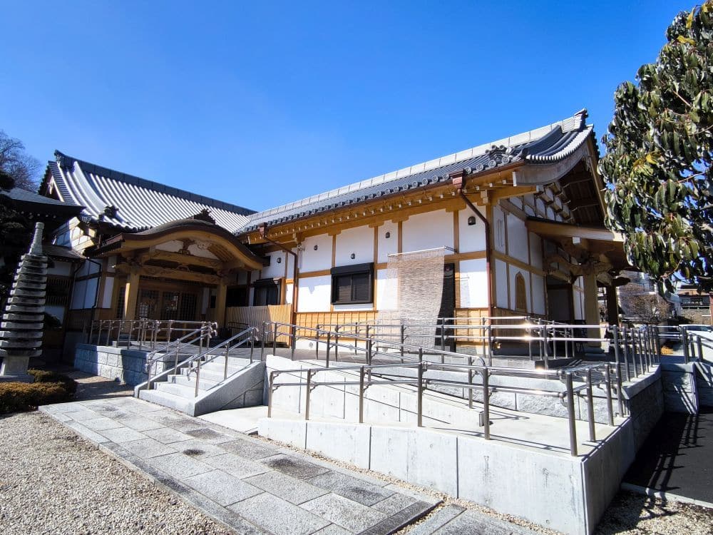 Der Hosenji-Tempel in Hino ist ein traditionelles japanisches Gebäude mit Ziegeldach und hölzernen Akzenten unter blauem Himmel, das über Rampen und Geländer zugänglich ist. Eine steinerne Pagode ziert die linke Seite, während sich auf der rechten Seite ein Baum erhebt, der seine reiche Geschichte widerspiegelt.
