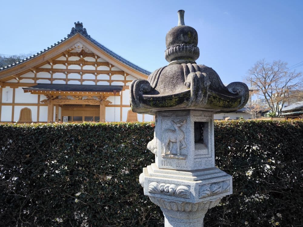 Eine traditionelle japanische Steinlaterne steht vor einem Holzgebäude mit Ziegeldach am Hosenji-Tempel in Hino, einem Ort der Ruhe. An der Laterne sind dekorative Schnitzereien zu sehen, die sich vor einem klaren blauen Himmel und einer gestutzten Hecke abzeichnen.