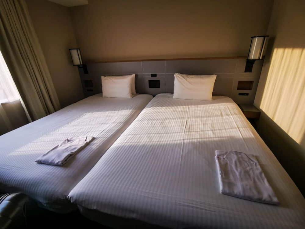 In einem Hotelzimmer stehen zwei Einzelbetten mit weißer Bettwäsche und Kissen, auf denen jeweils ein gefaltetes Tobu-Nachthemd oder ein Bademantel liegt. Warmes Sonnenlicht strömt durch das Fenster und wirft sanfte Schatten auf die Betten.