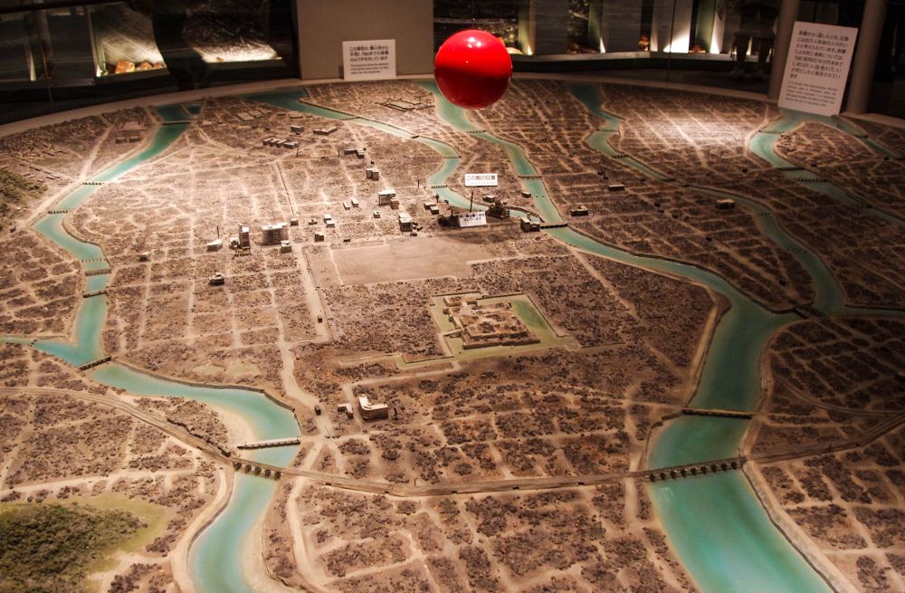 Ein detailliertes Diorama einer Stadt mit gewundenen blauen Flüssen und spärlichen Gebäuden zeigt eine große rote Kugel, die über dem Zentrum hängt und einen Ort des Friedens in der Stadt hervorhebt. Im Hintergrund sind Bildungsschilder zu sehen.