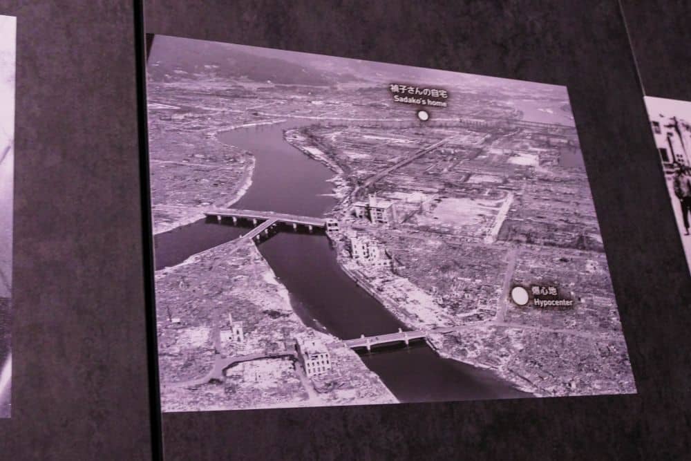 Ein schwarz-weißes Luftbild zeigt Hiroshima nach dem Atombombenabwurf, mit weitreichender Zerstörung, Brücken über einen Fluss und beschrifteten Punkten, die Sadakos Haus, das Hypocenter und Erinnerungen an eine dem Frieden gewidmete Zukunft markieren.