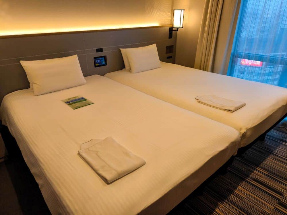 Zwei sauber gemachte Einzelbetten mit weißer Bettwäsche und Kissen stehen in diesem Tobu-Hotelzimmer nebeneinander. Auf jedem Bett liegt ein gefalteter Schlafanzug, und ein Fenster mit blauen Vorhängen lässt natürliches Licht herein.