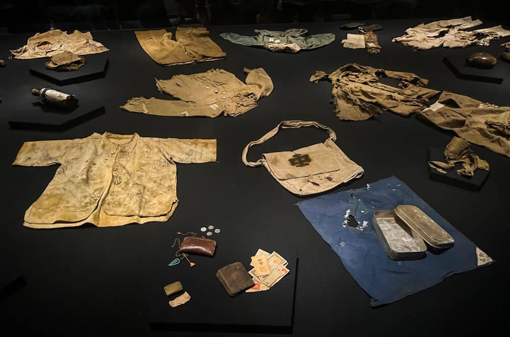 Verschiedene abgenutzte und beschädigte Kleidungsstücke, Taschen, Münzen und Behälter werden auf einer dunklen Fläche als historische Artefakte ausgestellt. Ihr gealtertes und beflecktes Aussehen erinnert an Geschichten vom Überleben und der anhaltenden Hoffnung auf Frieden in einer früheren Ära.