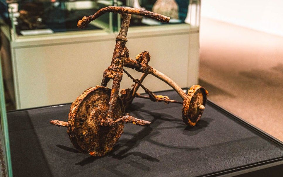 Ein stark verrostetes und korrodiertes Dreirad, das den verlorenen Frieden symbolisiert, steht auf einer schwarzen Plattform in einer Museumsumgebung, in der im Hintergrund Vitrinen und Ausstellungsstücke zu sehen sind.