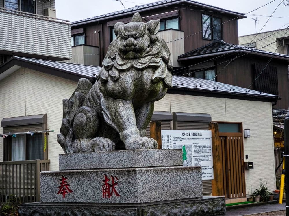 Eine steinerne Löwen-Hund-Statue (komainu) sitzt auf einem Granitsockel mit roten japanischen Schriftzeichen und bewacht den Eingang zu einem Hikawa-Schrein vor einem modernen Gebäude in einem japanischen Wohnviertel.