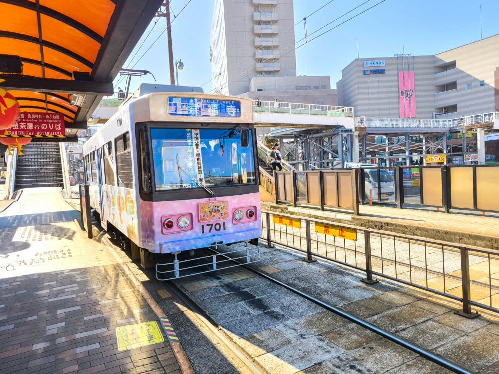 Eine bunte Straßenbahn mit der Nummer 1701 wartet an einer sonnigen Außenstation in Nagasaki, umgeben von Gebäuden und hellen Schildern. Der Bahnsteig wird von einem gelb-orangefarbenen Vordach beschattet - perfekt für alle, die Tram-Tipps suchen oder mit der Tram Nagasaki fahren wollen.