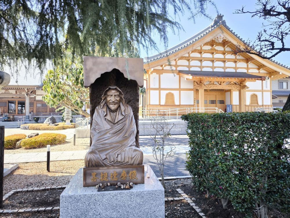 Eine Bronzestatue eines sitzenden älteren Mannes mit glücklichem Gesichtsausdruck ziert den Hosenji-Tempel in Hino, der auf einem Steinsockel ruht. Sträucher und Bäume umrahmen diesen Ort der Ruhe vor dem traditionellen japanischen Holztempel unter einem klaren Himmel.