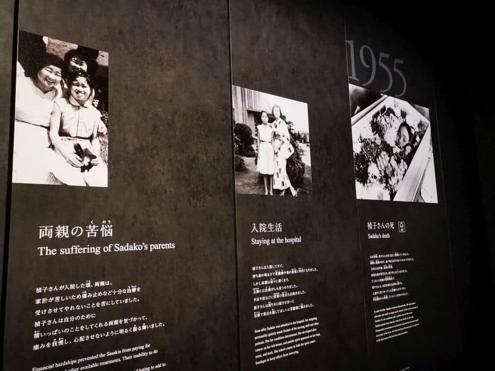 Drei schwarze Tafeln zeigen historische Fotos und Text. Die linke zeigt eine lächelnde Familie, die mittlere ein Mädchen mit einer Frau im Freien und die rechte ein junges Mädchen in einem Sarg. Die Tafeln erzählen von Sadakos Leben, ihrem Leiden im Jahr 1955 und ihrem Wunsch nach Frieden.
