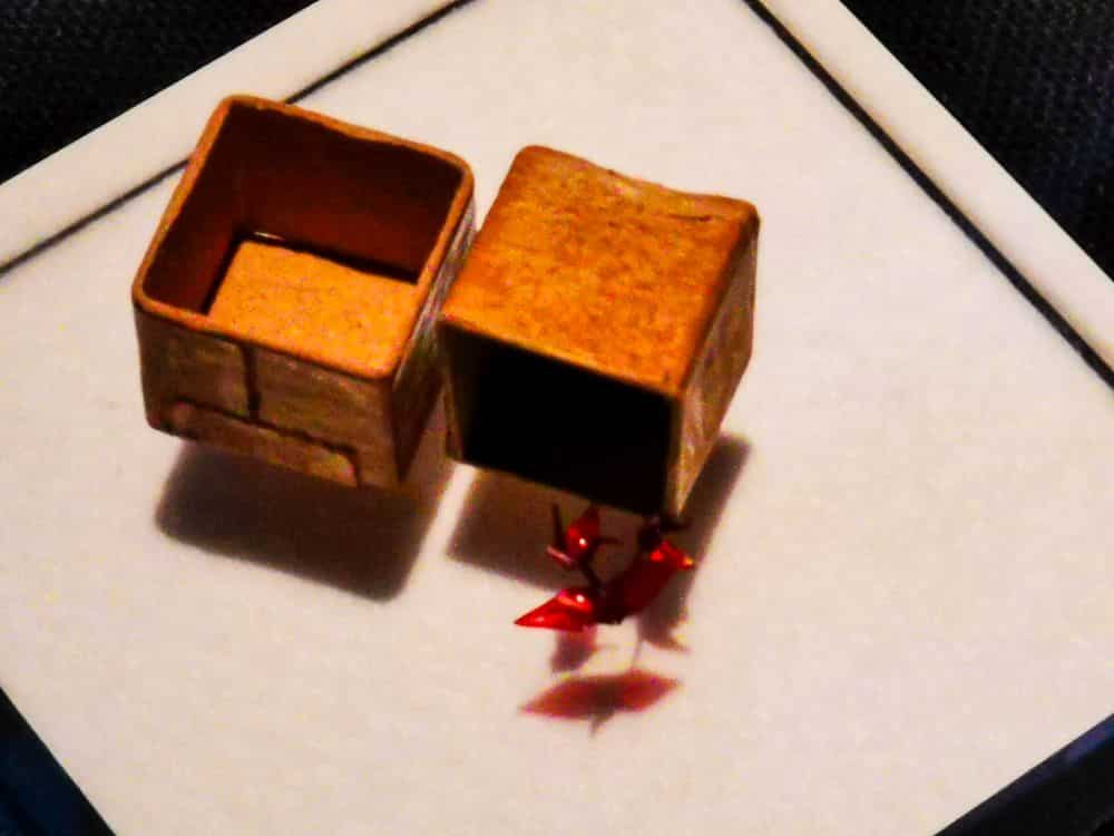 Auf einer weißen Fläche steht eine kleine, offene, goldfarbene Schachtel, deren Deckel hochgeklappt ist. Unter der Schachtel befinden sich zwei winzige, rot glänzende Origami-Kraniche - Symbole für Frieden und zarte Schönheit.