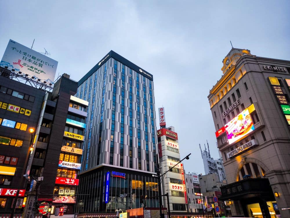 Stadtbild in der Abenddämmerung mit modernen und älteren Gebäuden, darunter auch Tobu-Filialen, mit bunten Leuchtreklamen und Werbetafeln in japanischer Sprache unter einem bewölkten Himmel. Belebte städtische Atmosphäre mit lebhafter kommerzieller Aktivität.