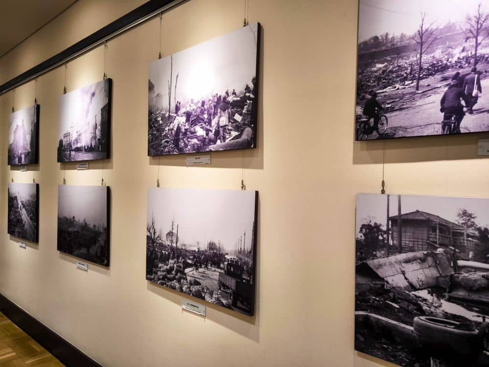 Historische Schwarz-Weiß-Fotos, die auf einer Galeriewand im Memorial Museum ausgestellt sind und Szenen des Großen Kanto-Erdbebens zeigen, darunter Menschen, zerstörte Gebäude und Trümmer. Die Fotos sind in zwei Reihen aufgehängt, unter denen sich Beschriftungen befinden.