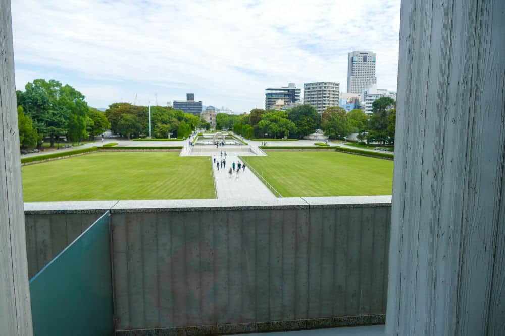 Blick auf eine weite, grüne Wiese und einen Weg mit Menschen, die in Ruhe spazieren gehen, eingerahmt von zwei hohen Säulen; im Hintergrund erscheinen Stadtgebäude und Bäume unter einem teilweise bewölkten Himmel.