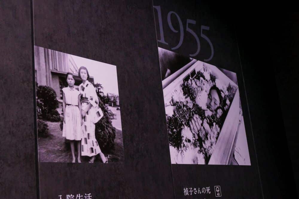 An einer dunklen Wand unter dem großen Text 1955 sind zwei Schwarz-Weiß-Fotos zu sehen. Das linke Foto zeigt zwei Frauen, die im Freien stehen; das rechte zeigt eine Person, die in einem Sarg liegt, umgeben von Blumen - ein düsterer Moment, der an Frieden erinnert.
