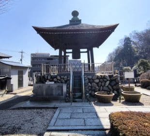 Ein großer traditioneller japanischer Glockenturm steht im Innenhof des Hosenji, umgeben von Steinwegen und angelegten Büschen, mit Gebäuden und Bäumen, die an einem sonnigen Tag im Hintergrund zu sehen sind. Joya no Kane