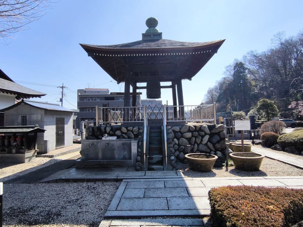 Der Hosenji-Tempel in Hino ist ein traditioneller japanischer Glockenturm auf einer steinernen Plattform mit einer Treppe nach oben, Stein- und Kieswegen und Bäumen - ein Ort der Ruhe unter dem klaren blauen Himmel inmitten der umliegenden Gebäude.