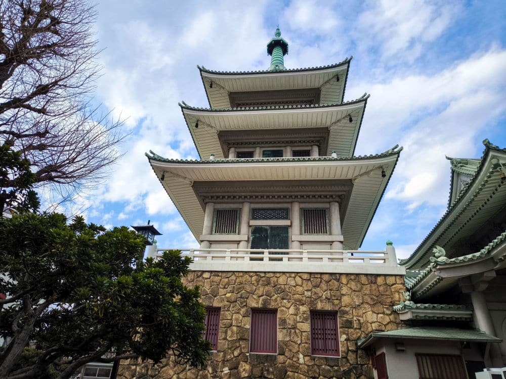 Eine traditionelle mehrstöckige Pagode mit grünen Ziegeldächern und einem Steinsockel steht vor einem teilweise bewölkten Himmel, eingerahmt von Bäumen auf der linken Seite - eine eindrucksvolle Erinnerung an die Geschichte in der Nähe des Great Kanto Earthquake Memorial Museum.