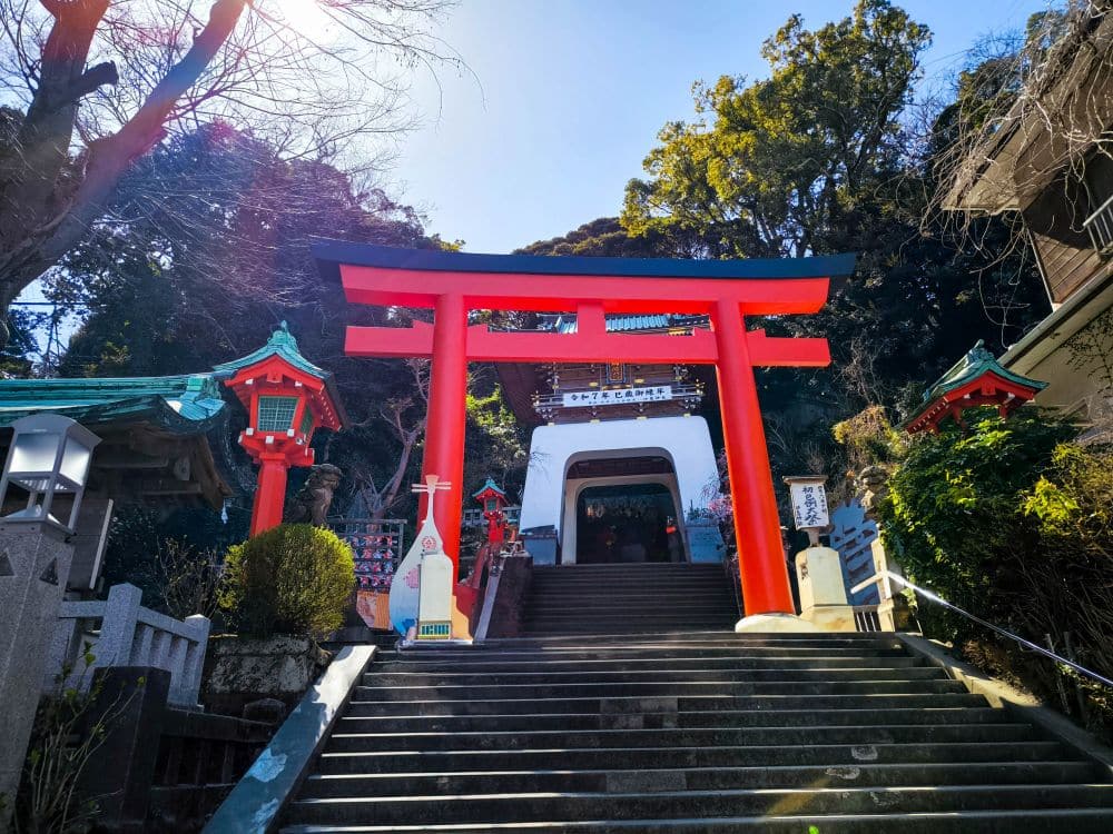 Ein großes rotes Torii-Tor steht an der Spitze einer Steintreppe, die zum Hetsunomiya-Schrein führt, umgeben von Bäumen und traditionellen Laternen unter einem strahlend blauen Himmel.