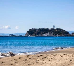 Ein Sandstrand mit sanften Wellen im Vordergrund und die Insel Enoshima in der Ferne, mit einem Leuchtturm und Gebäuden unter einem klaren blauen Himmel.