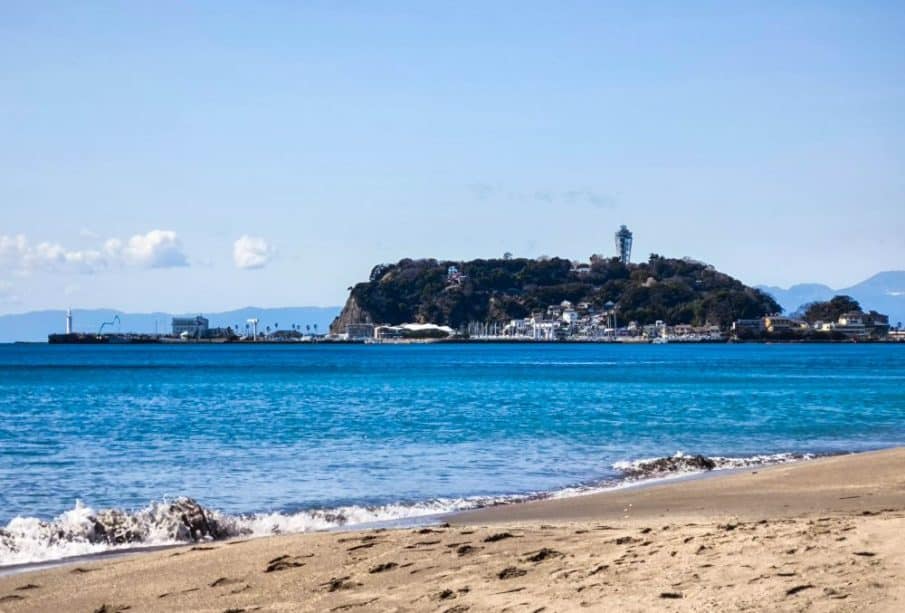 Ein Sandstrand mit sanften Wellen im Vordergrund und die Insel Enoshima in der Ferne, mit einem Leuchtturm und Gebäuden unter einem klaren blauen Himmel.