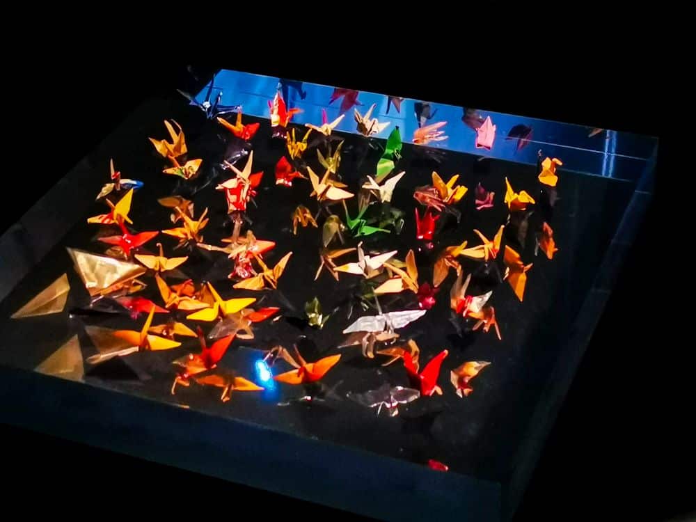 Auf einer schwarzen, glasähnlichen Fläche sind bunte Origami-Kraniche angeordnet, die den Frieden symbolisieren und vor einem dunklen Hintergrund dramatisch beleuchtet werden.
