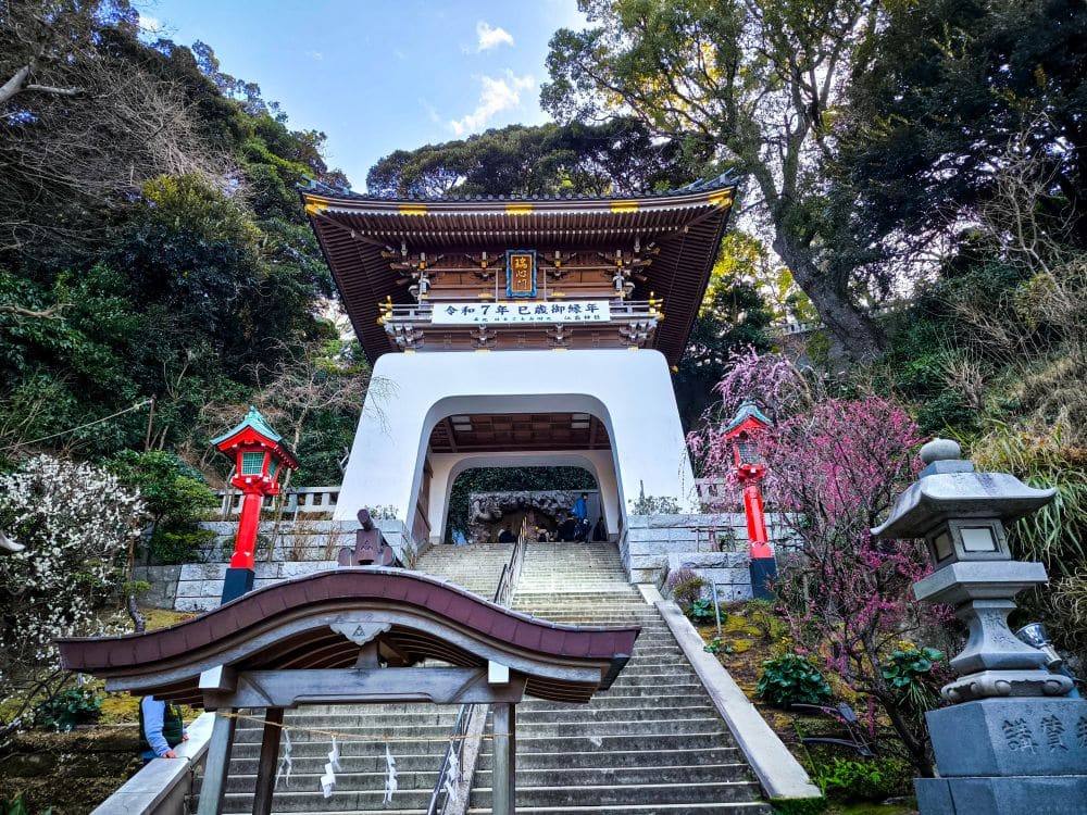 Steinstufen führen hinauf zum Tor des Hetsunomiya-Tempels, das von roten Laternen, blühenden Bäumen und üppigem Grün umrahmt ist, mit einer Holzkonstruktion am Fuß der Treppe.