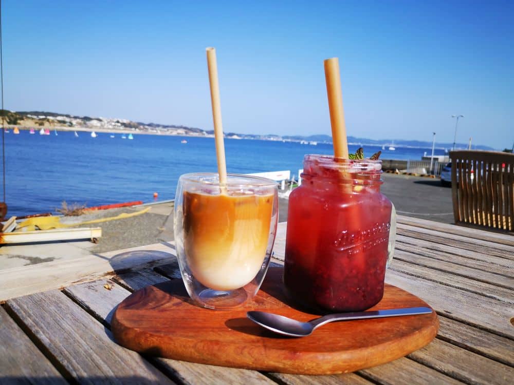 Zwei kalte Getränke stehen auf einem Holzbrett auf einem Tisch im Freien am Meer in Enoshima: ein eisgekühlter Milchkaffee in einem Glas und ein rotes Beerengetränk in einem Einmachglas. Im Hintergrund sind ruhiges blaues Wasser, Boote und ein klarer Himmel zu sehen.